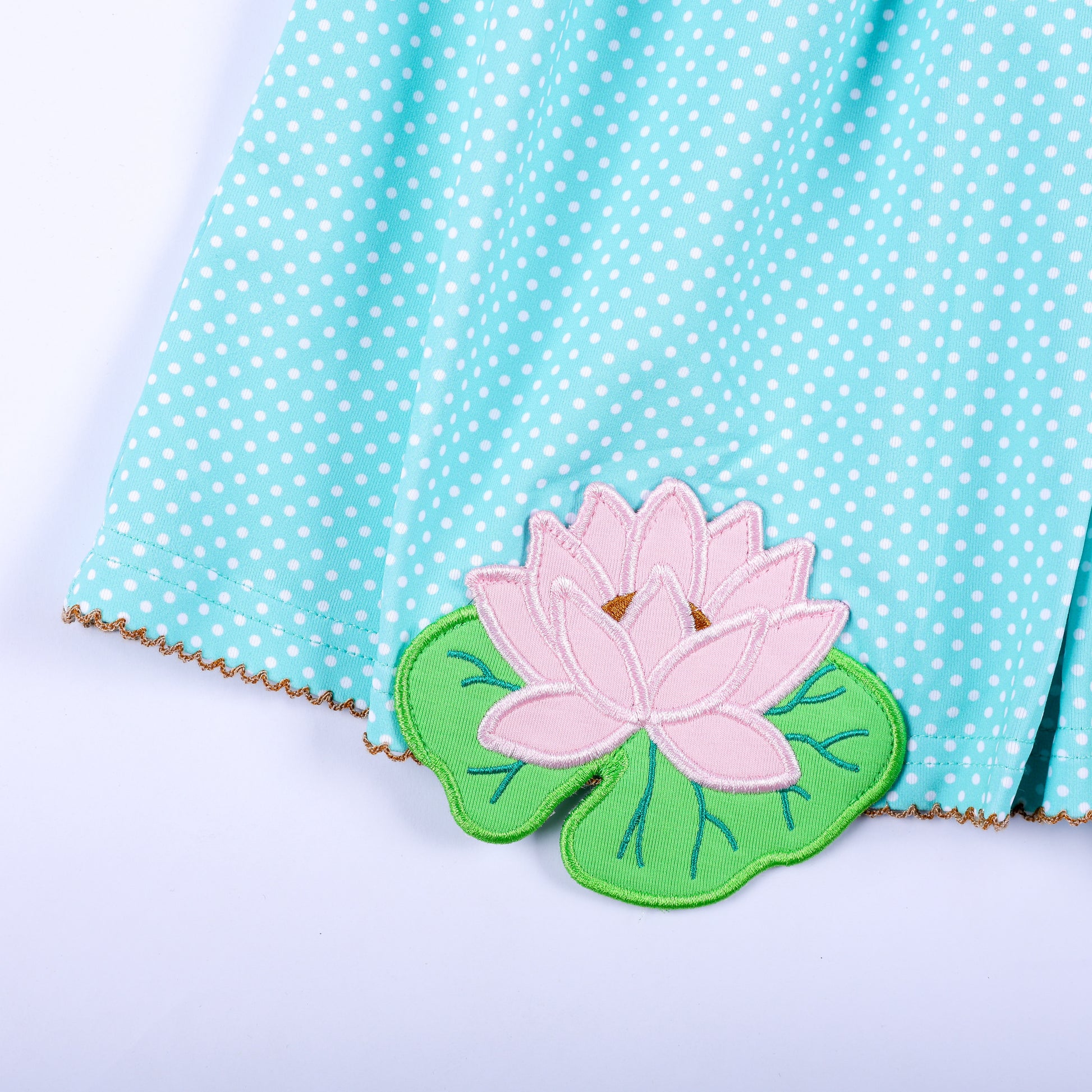 Close-up view of pink lotus flower applique hand sewn on hem of aqua polka dot smocked dress for girls.