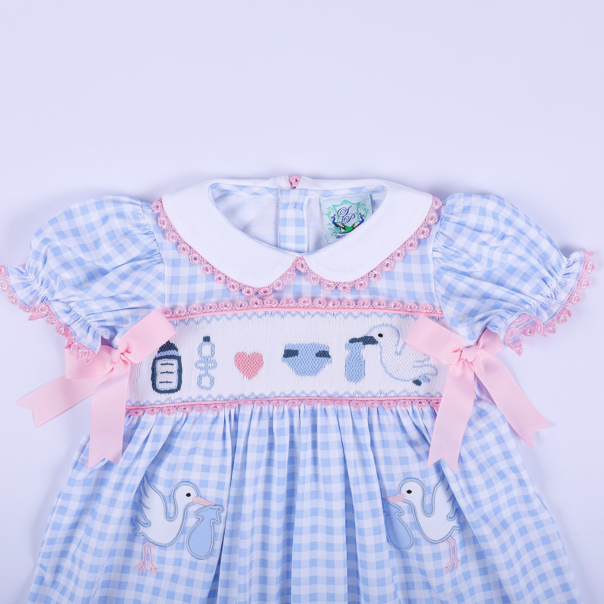 Mid shot of hand smocked baby shower announcement dress showing embroidered motifs, pink bows, and Peter Pan collar.