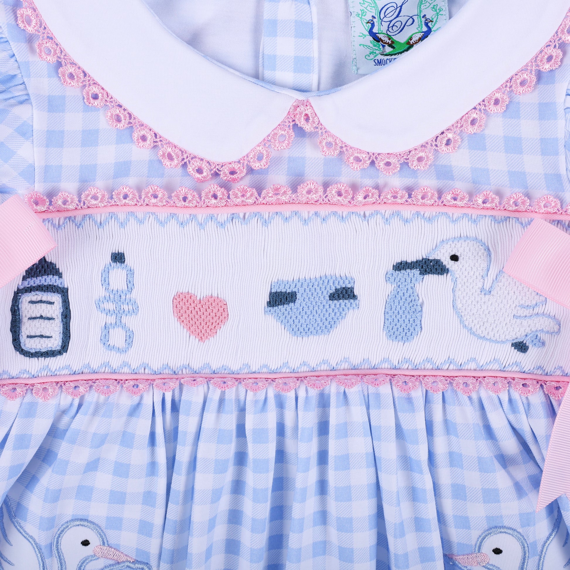 Flat lay close up of hand embroidered baby motifs including bottle, rattle, heart, diaper, and stork on smocked dress.