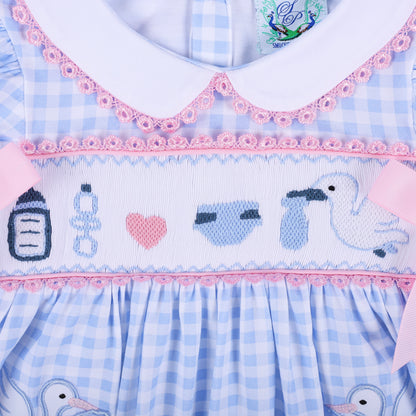 Flat lay close up of hand embroidered baby motifs including bottle, rattle, heart, diaper, and stork on smocked dress.