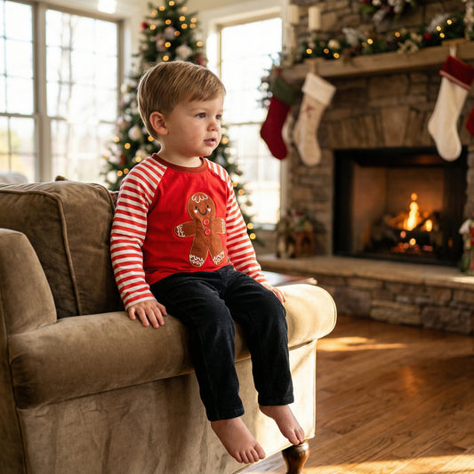 Toddler boy wearing red gingerbread Christmas shirt with striped sleeves sitting by a fireplace.