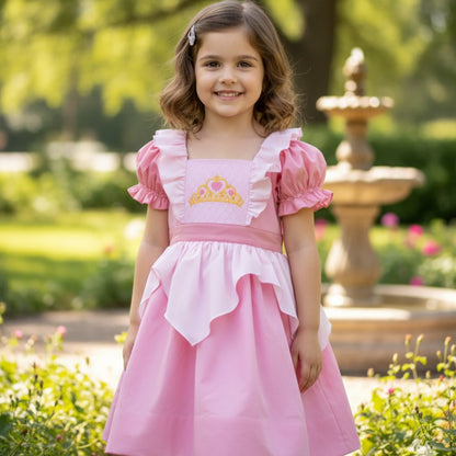 Smiling little girl in a garden wearing a pink princess dress with an embroidered gold crown and puff sleeves.