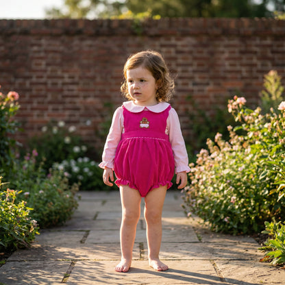 Toddler girl wearing a pink corduroy smocked bubble romper with a gingerbread applique and long sleeve shirt in a garden.