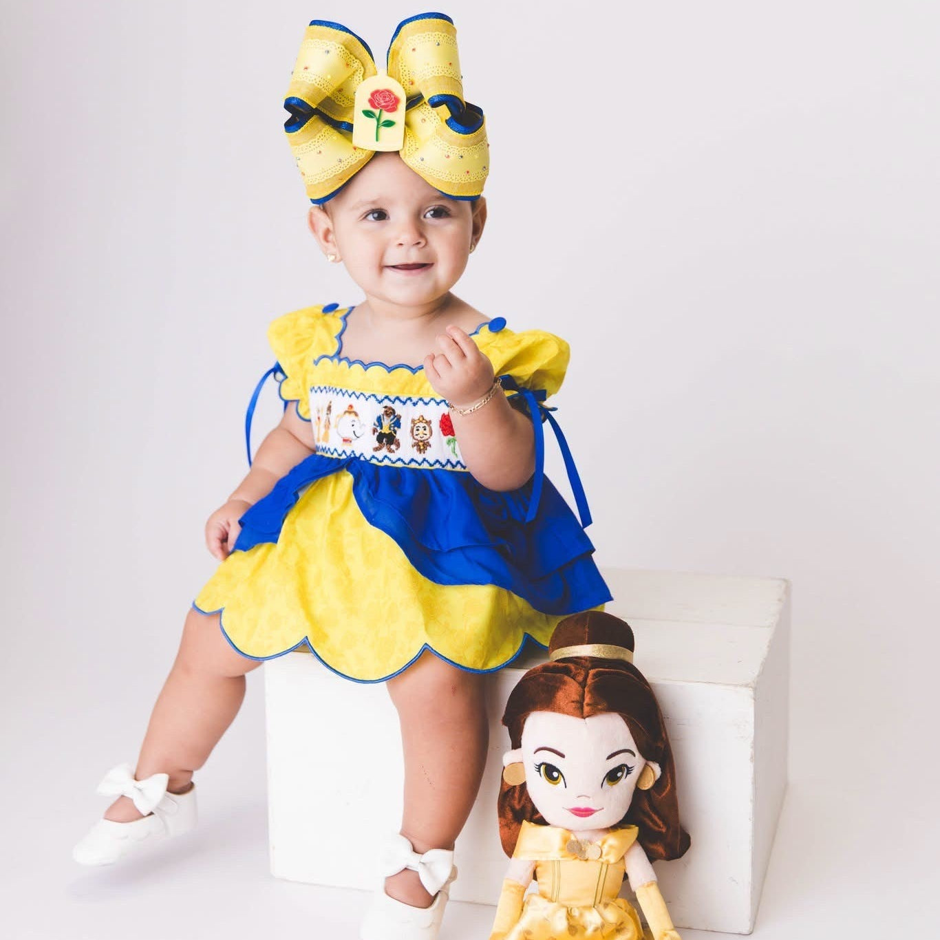 Toddler wearing a yellow and blue custom smocked Belle bloomer set with a matching bow next to a doll