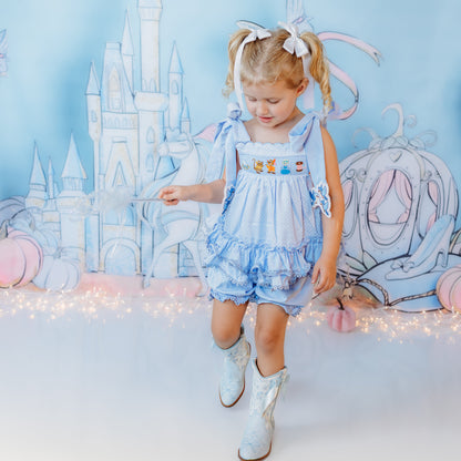 Toddler girl wearing a light blue hand-smocked Cinderella ruffle top and matching bloomer set with white boots in a fairy-tale castle photo set.