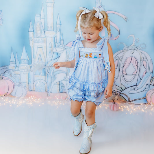 Toddler girl wearing a light blue hand-smocked Cinderella ruffle top and matching bloomer set with white boots in a fairy-tale castle photo set.