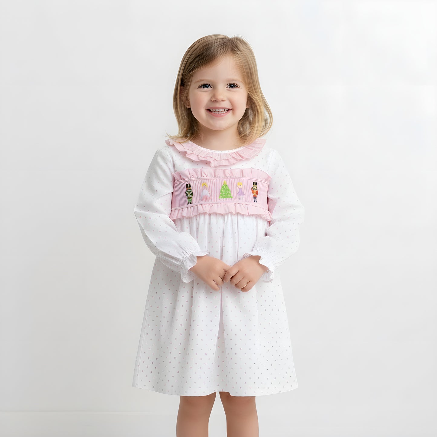 Smiling young girl model wearing a pink Nutcracker smocked Christmas dress against a white studio background.