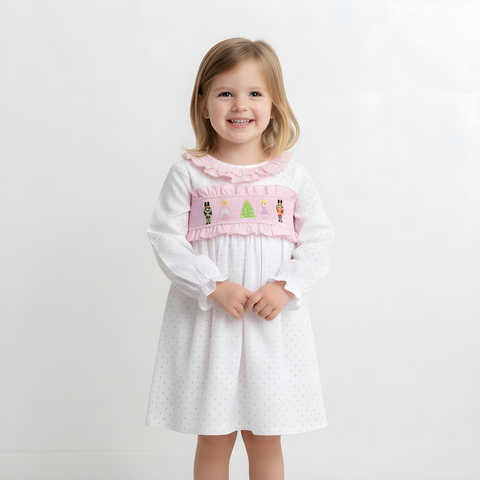 Smiling young girl model wearing a pink Nutcracker smocked Christmas dress against a white studio background.