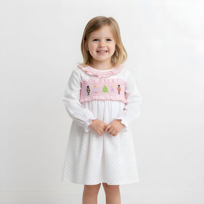 Smiling young girl model wearing a pink Nutcracker smocked Christmas dress against a white studio background.