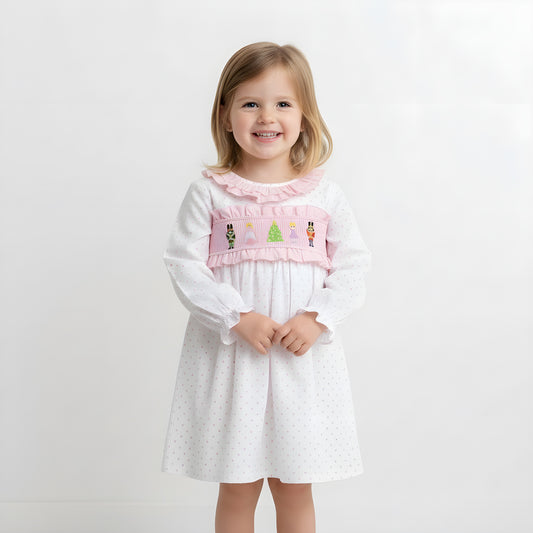 Smiling young girl model wearing a pink Nutcracker smocked Christmas dress against a white studio background.