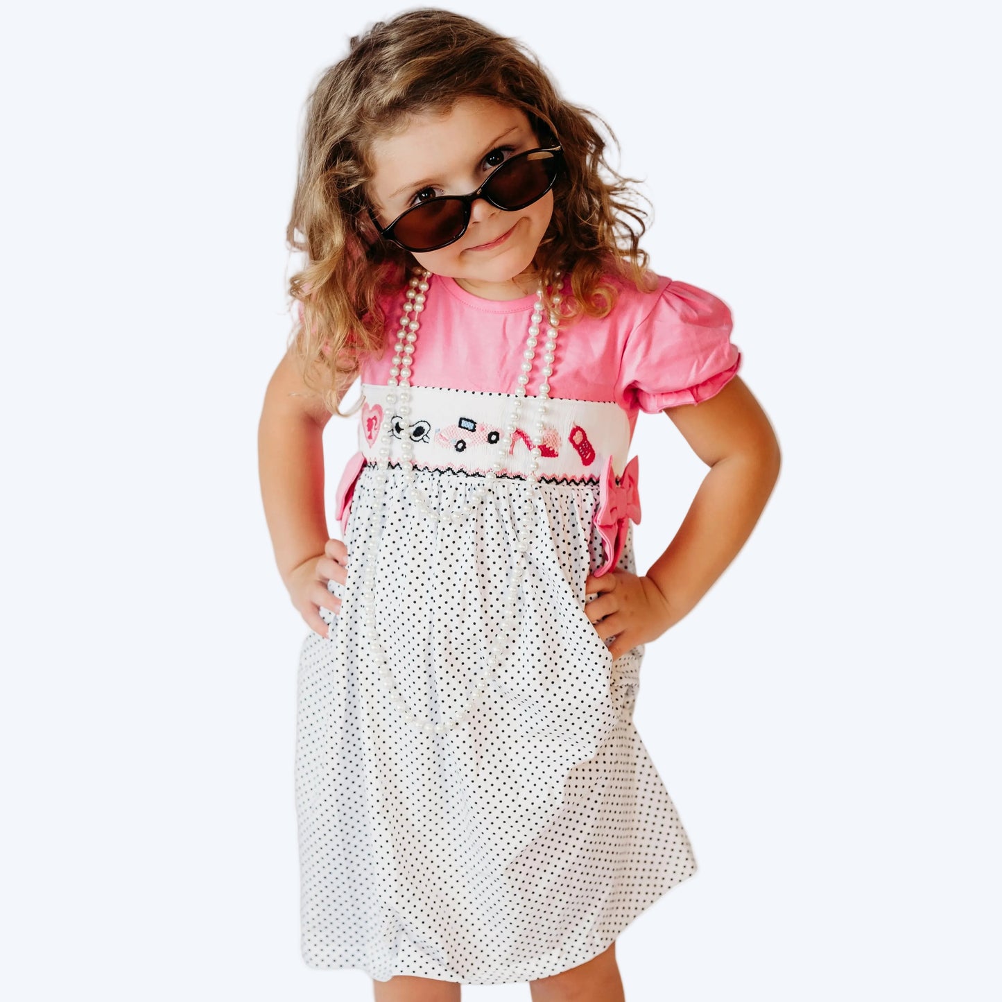 Young girl posing with sunglasses and pearls wearing a pink and white polka dot hand-smocked dress with fashion embroidery.