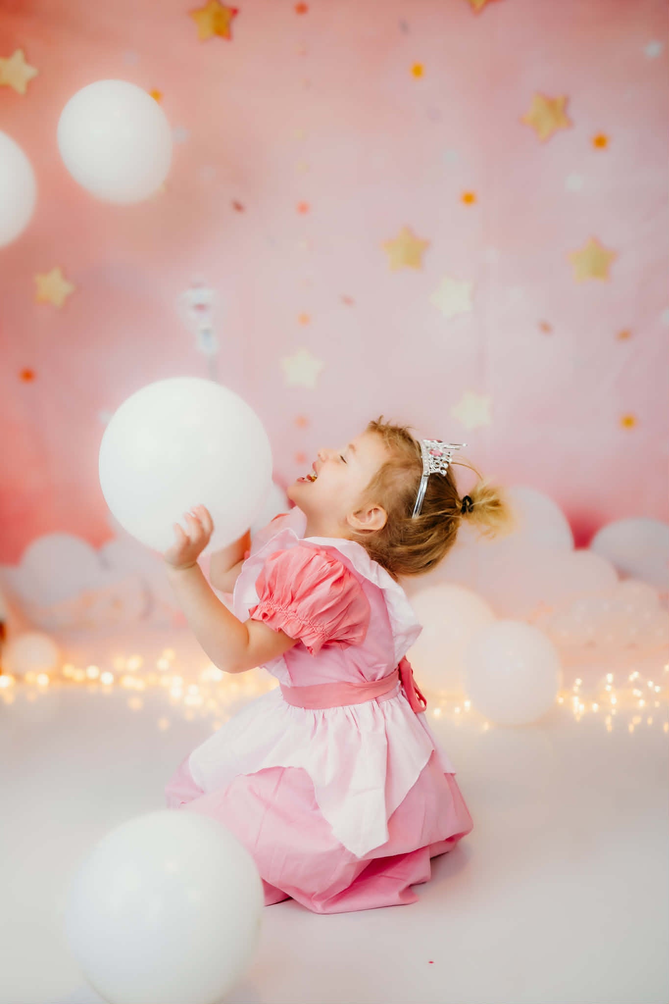 Toddler girl laughing in a pink puff sleeve princess birthday dress with a white apron overlay, holding a balloon.