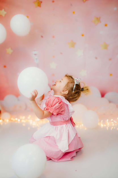 Toddler girl laughing in a pink puff sleeve princess birthday dress with a white apron overlay, holding a balloon.