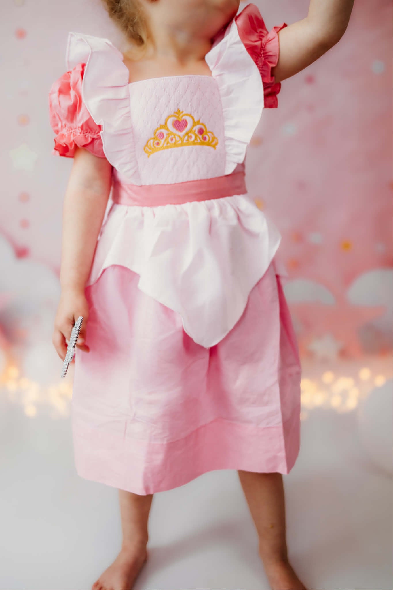 Toddler wearing a pink smocked princess dress with puff sleeves, white apron overlay, and embroidered crown.