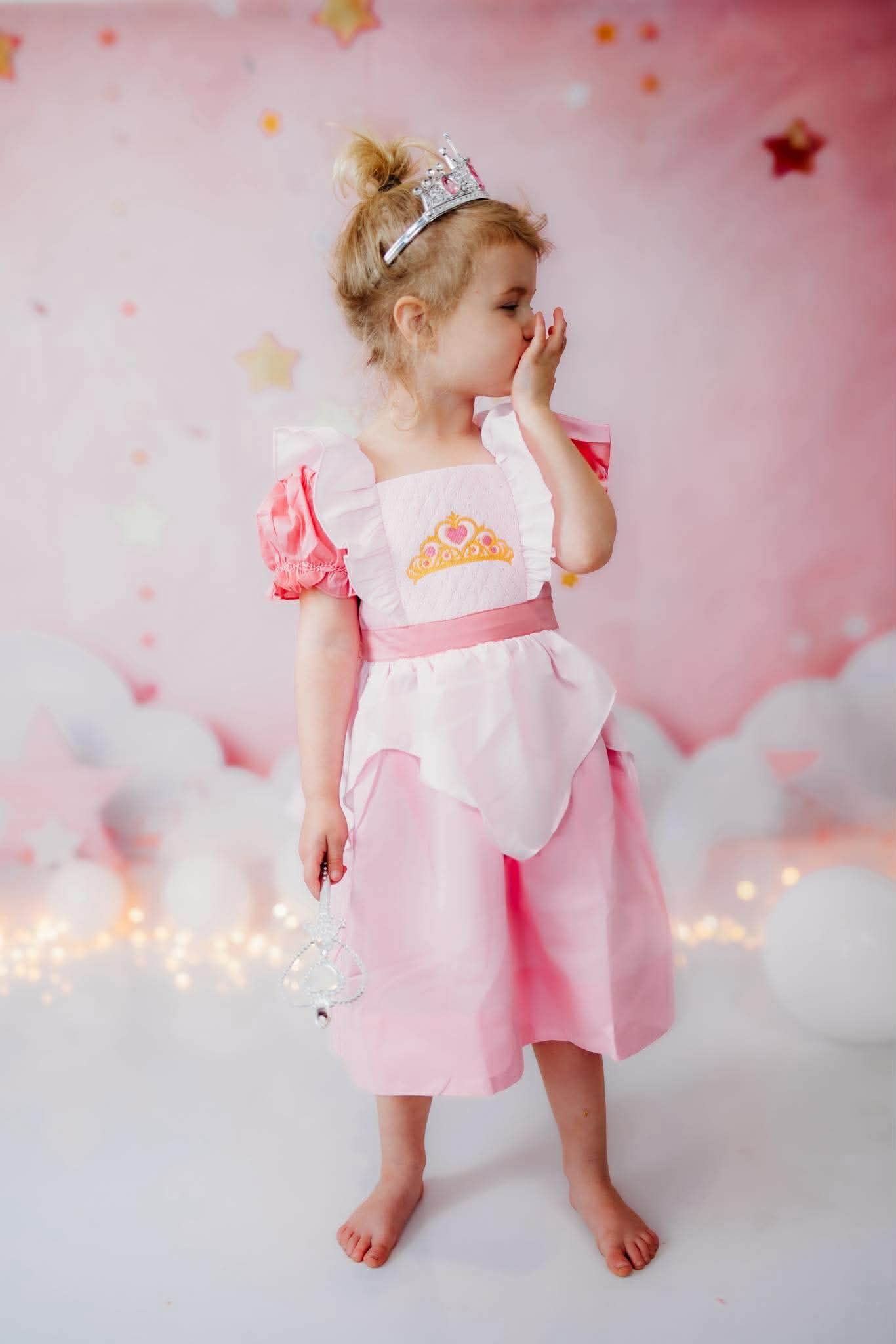 Toddler girl looking shy in a lovely smocked pink princess dress with ruffles and crown embroidery, holding a wand.
