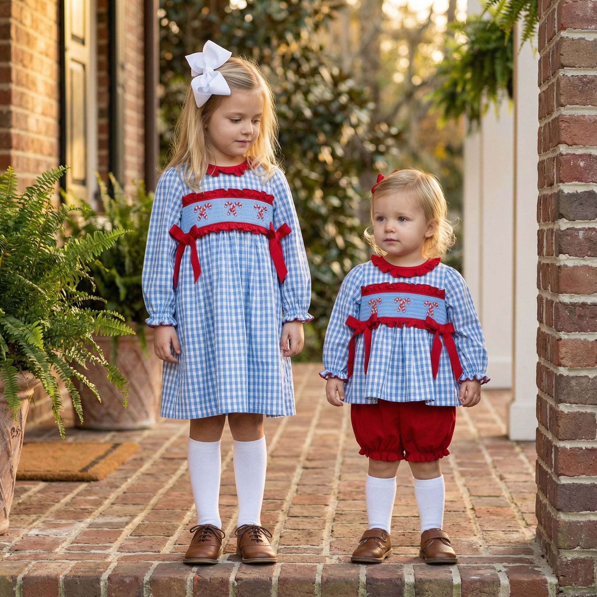 Sisters in matching blue gingham smocked Christmas outfits with red candy cane embroidery and red bows.