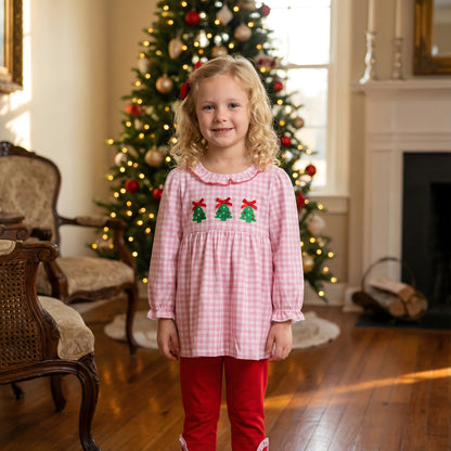 Girl in Embroidered Christmas Tree Trio Girls Pants Set, featuring a pink gingham smocked top and red pants, by a Christmas tree.