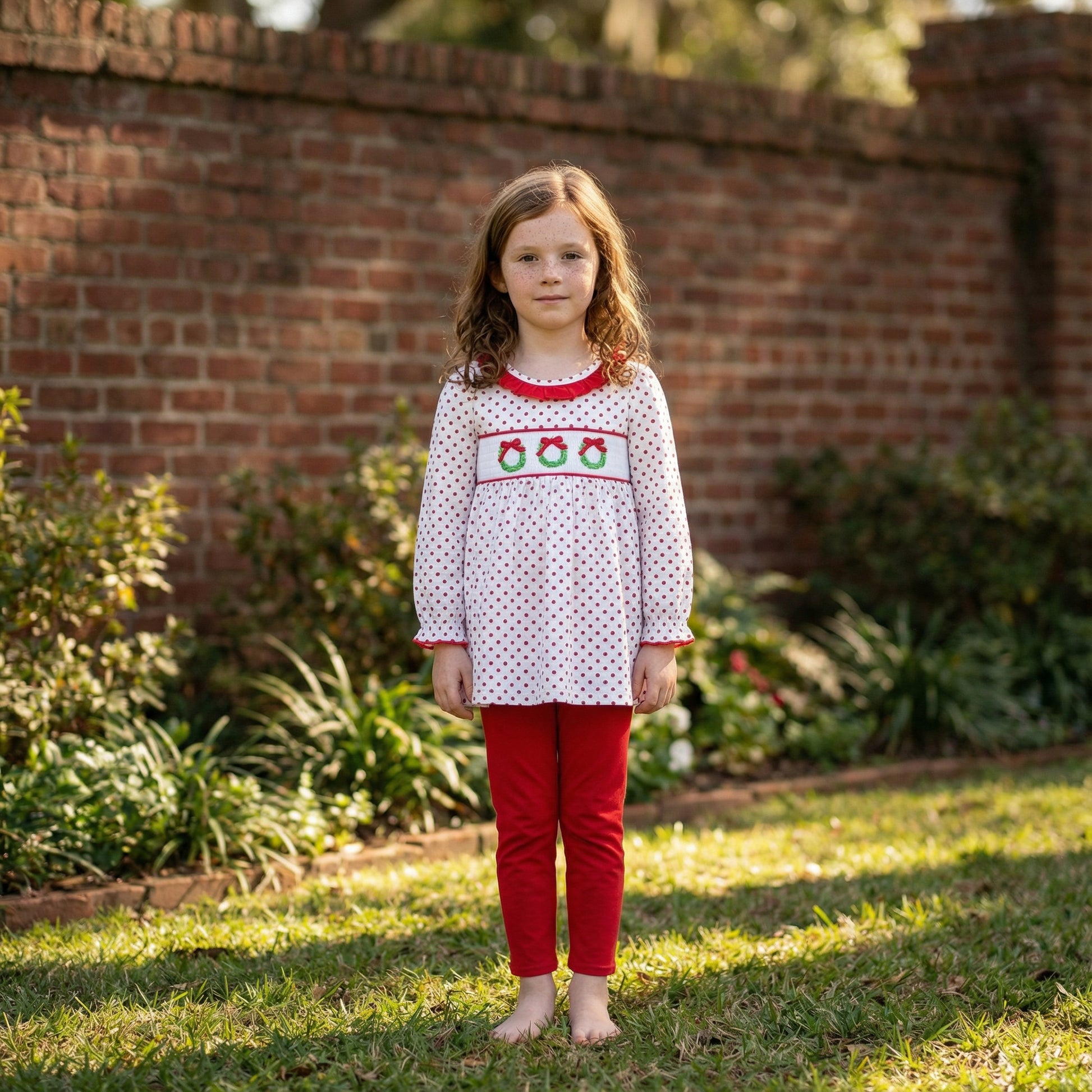 Girl in white smocked top with red polka dots and three Christmas wreaths, and red pants, standing outside.