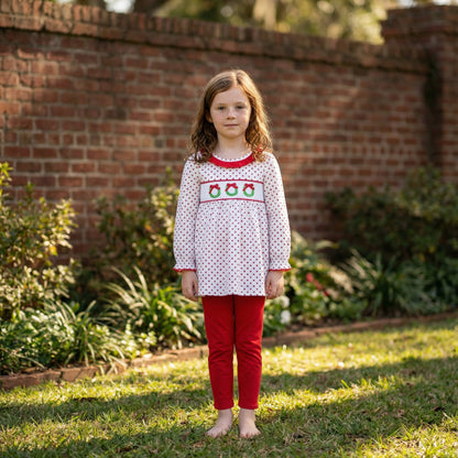Girl in white smocked top with red polka dots and three Christmas wreaths, and red pants, standing outside.