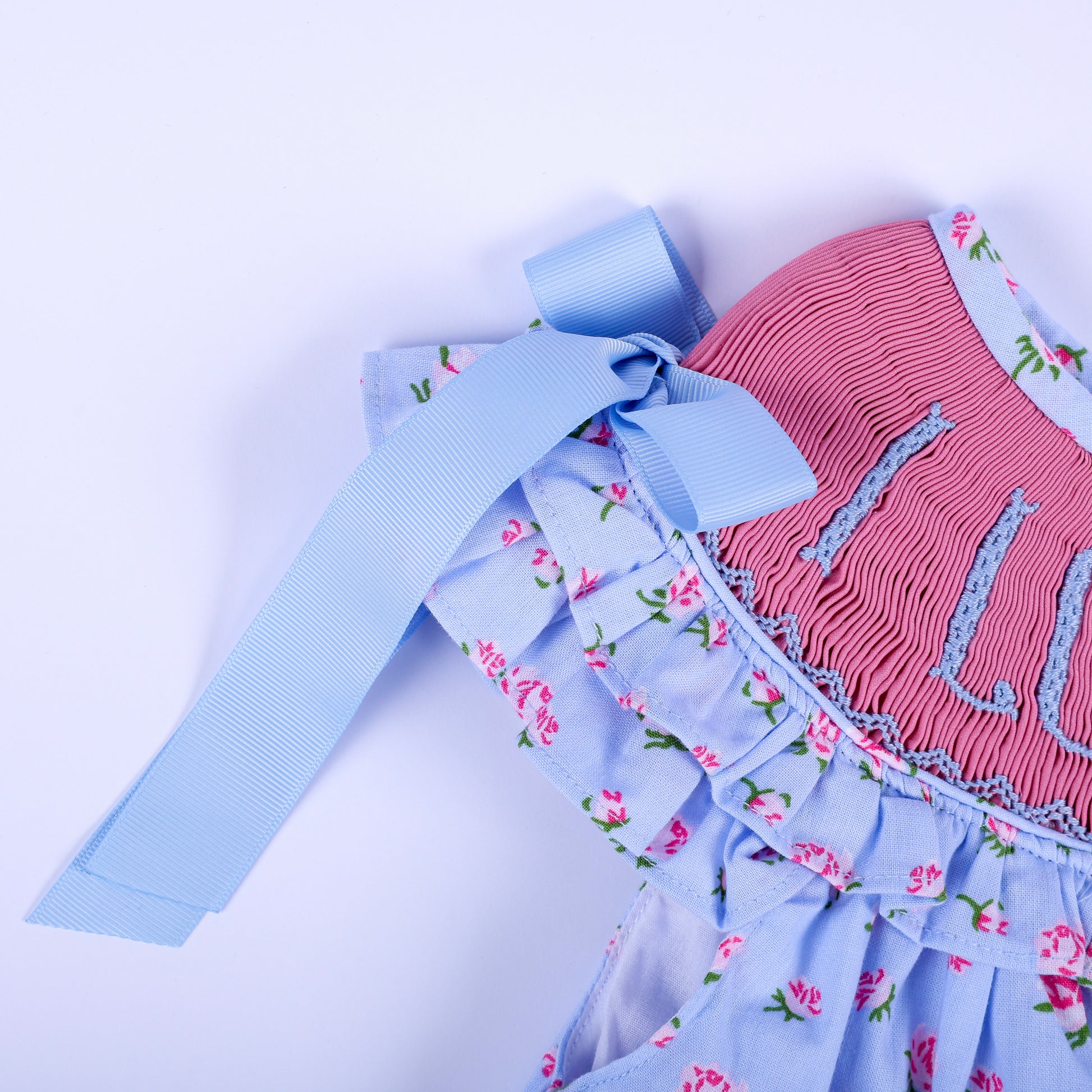 Detailed shot of light blue ribbon shoulder bow and ruffled sleeve on girls floral hand-smocked I Love Mom dress.
