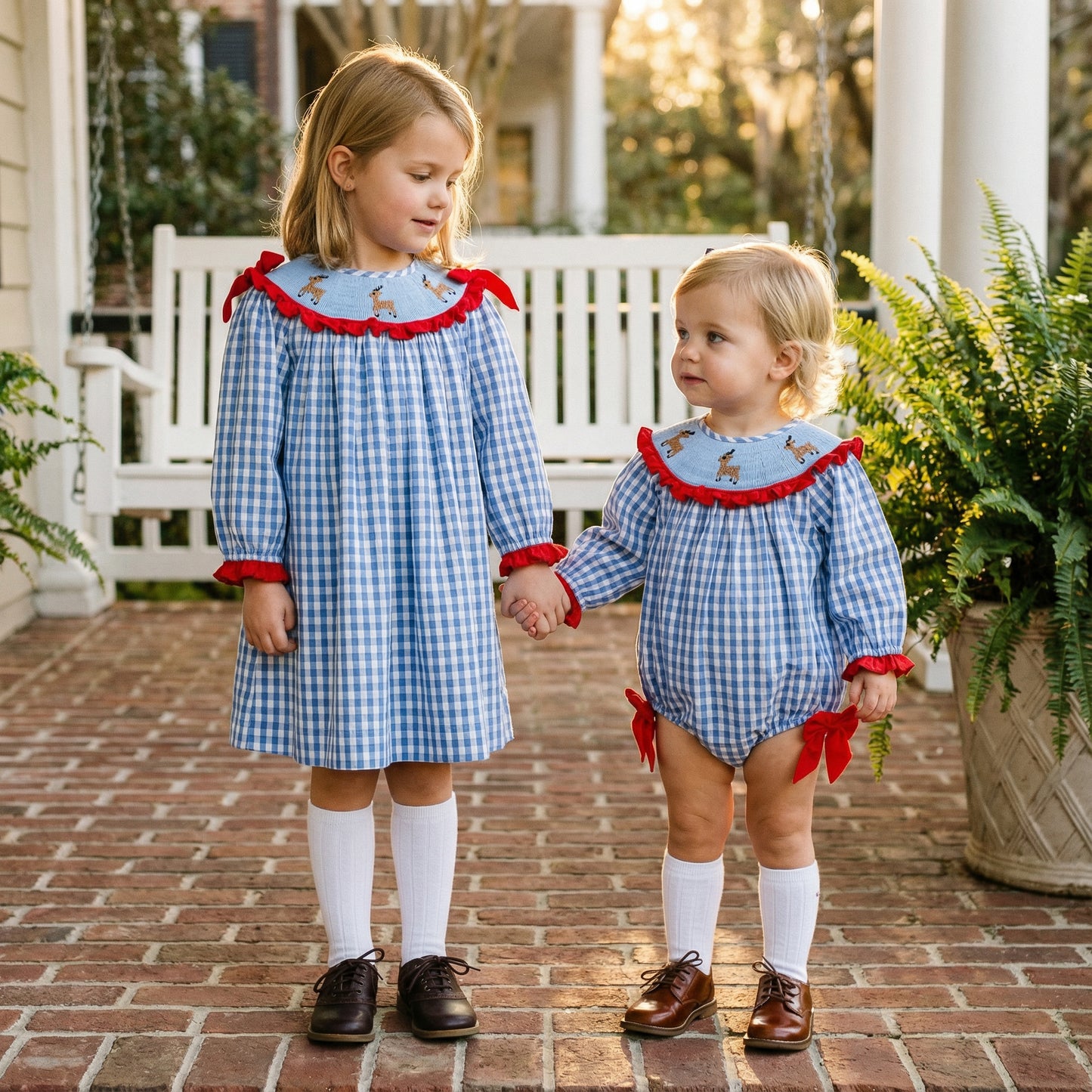 Two sisters holding hands in matching blue gingham outfits with hand-smocked red reindeer. Older girl in dress, younger in romper.