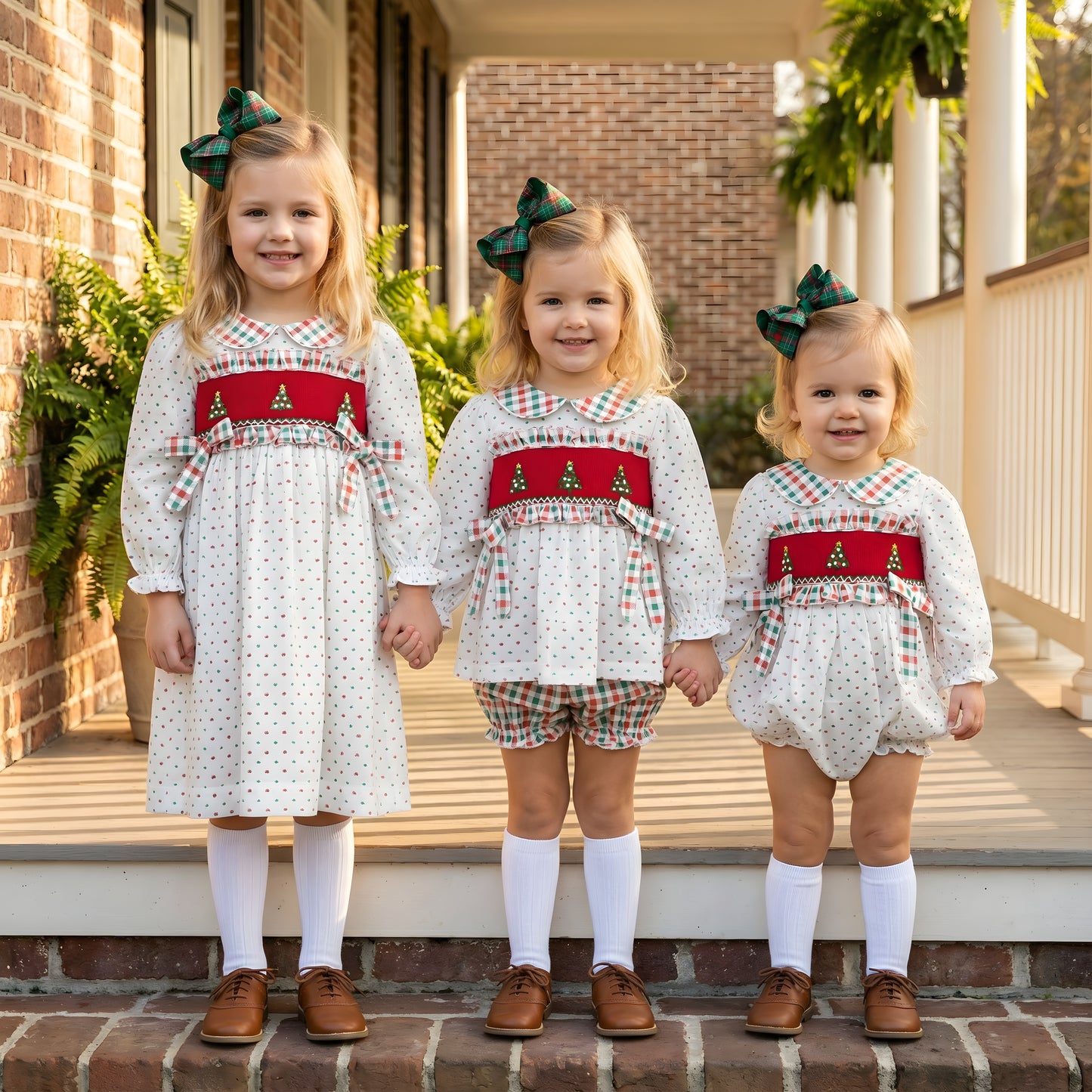 Three sisters in matching smocked Christmas outfits, featuring the Swiss Dot Christmas Tree Bloomer Set, dress, and a bubble.