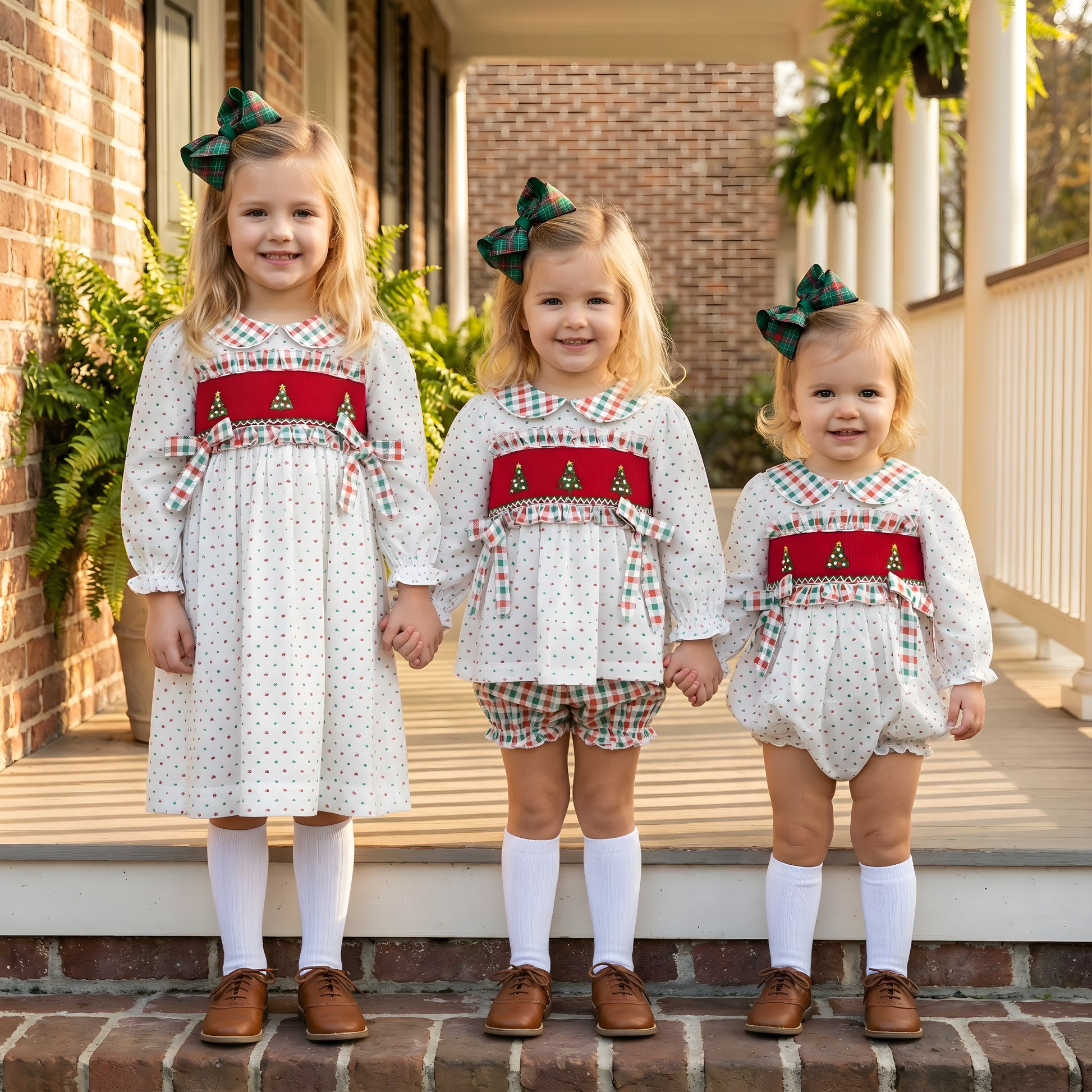 Three sisters in matching smocked Christmas outfits, featuring the Swiss Dot Christmas Tree Bloomer Set, dress, and a bubble.