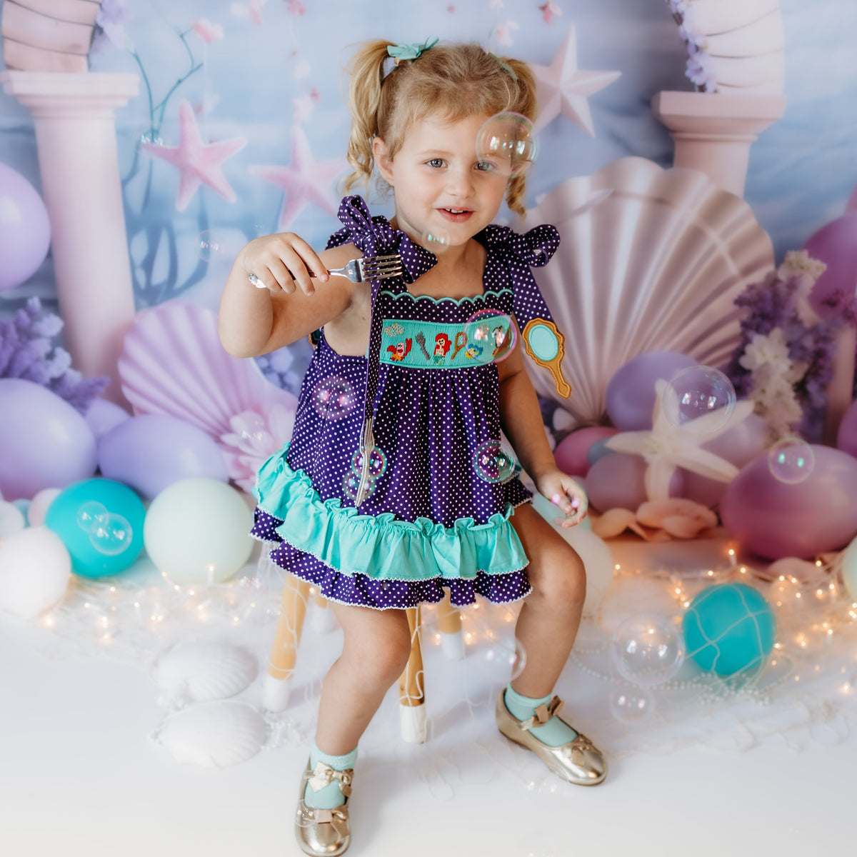 Toddler girl wearing a purple polka dot under the sea hand smocked bloomer set with turquoise ruffle trim in a mermaid-themed photoshoot.