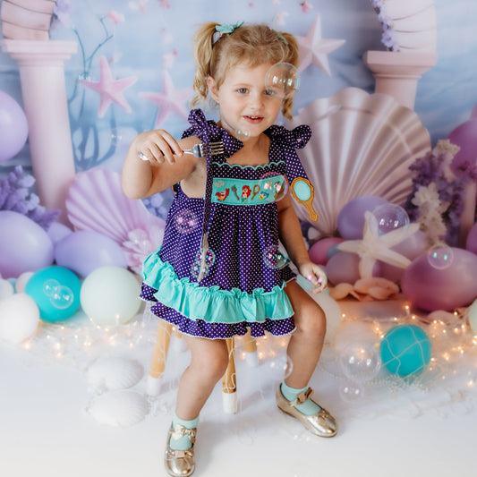 Toddler girl wearing a purple polka dot under the sea hand smocked bloomer set with turquoise ruffle trim in a mermaid-themed photoshoot.