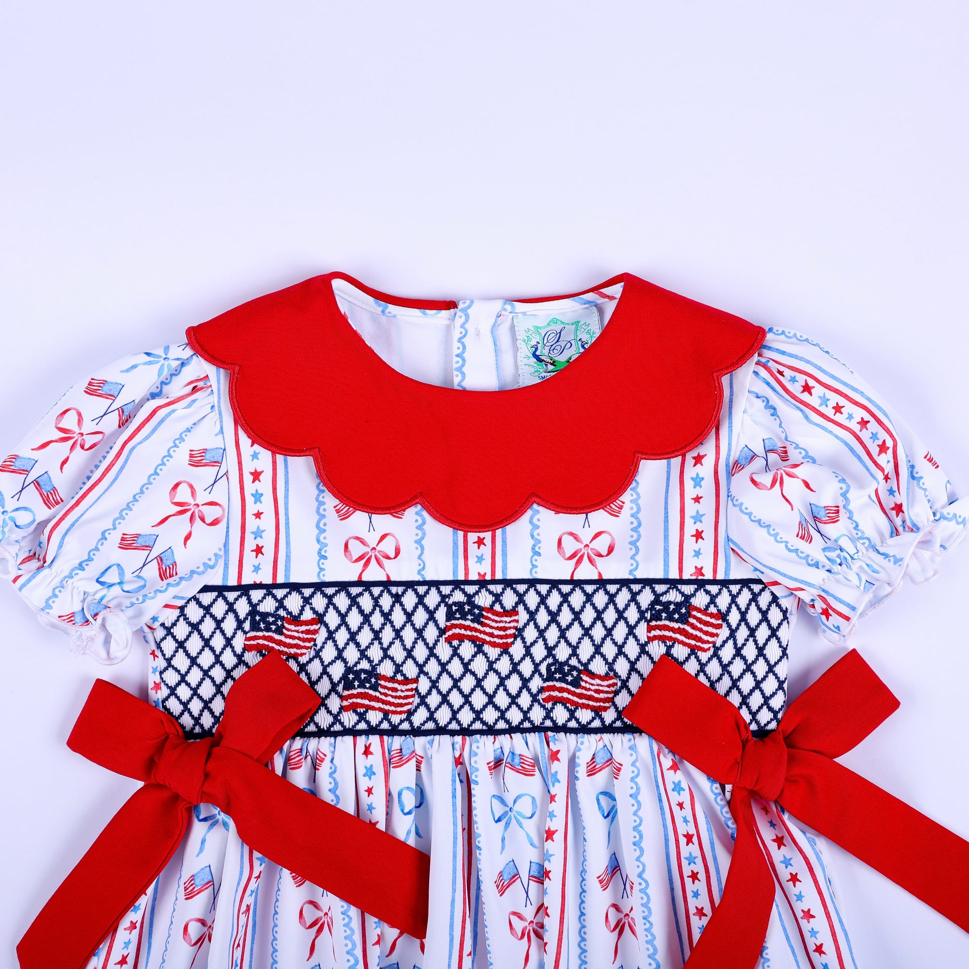 Close up of hand embroidered patriotic American flag smocking on a baby girl top with red scalloped collar and bows.