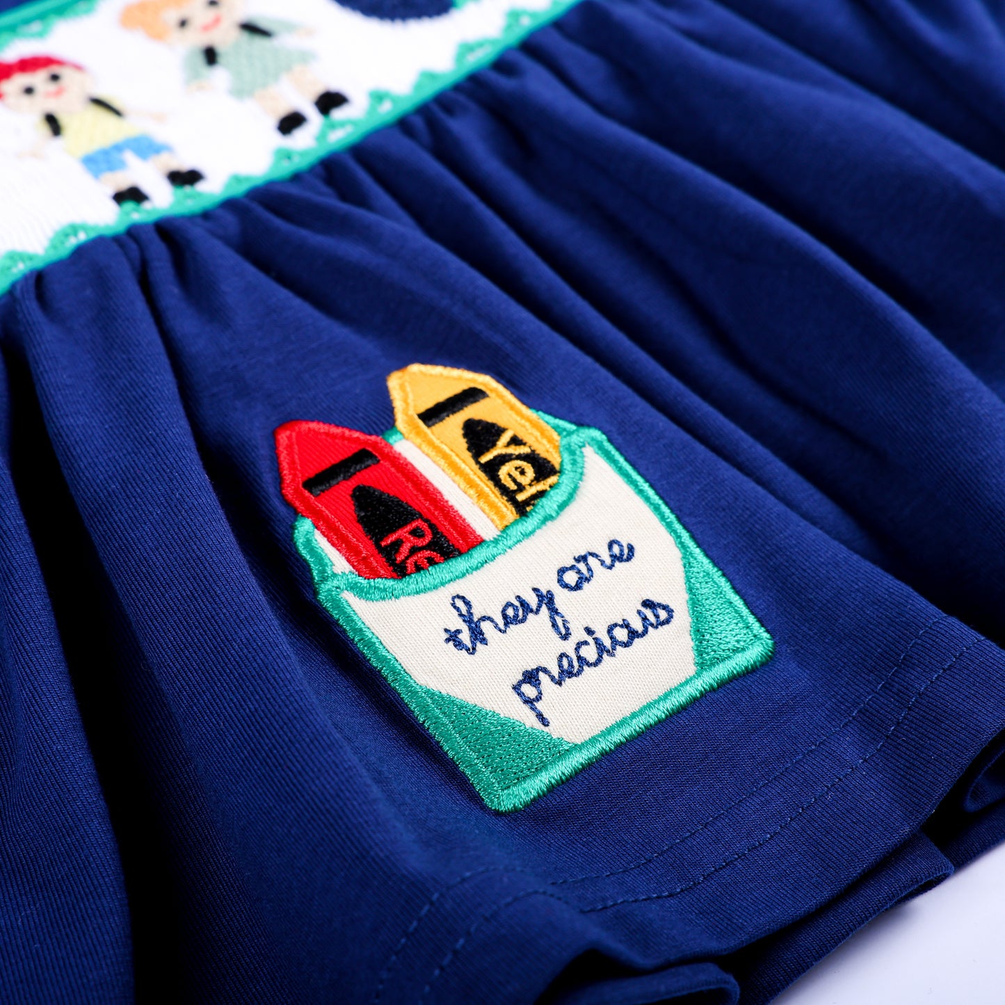 Close-up of hand embroidered crayon pocket with text reading they are precious on navy smocked top hem.