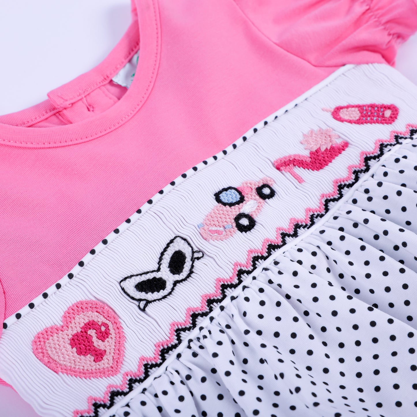 Macro shot of hand-smocked chest panel displaying pink hand-embroidered fashion accessories on polka dot bubble.