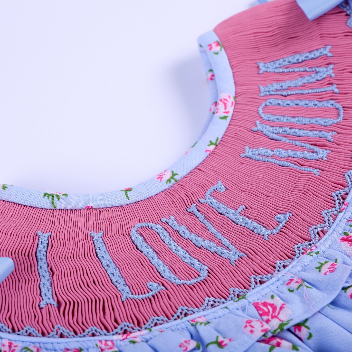Macro close-up showing intricate pink hand-smocked pleating and blue hand-embroidered I Love Mom text detail.