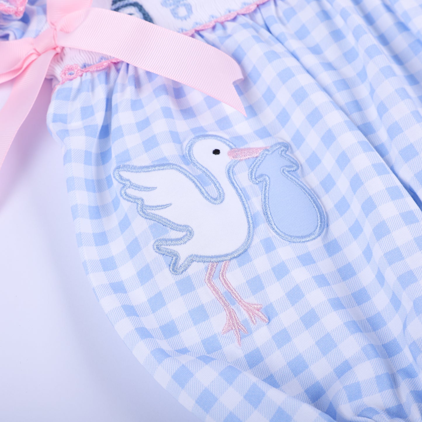 Detailed close-up of the hand-embroidered stork delivery motif on the light blue gingham gender reveal bubble romper.