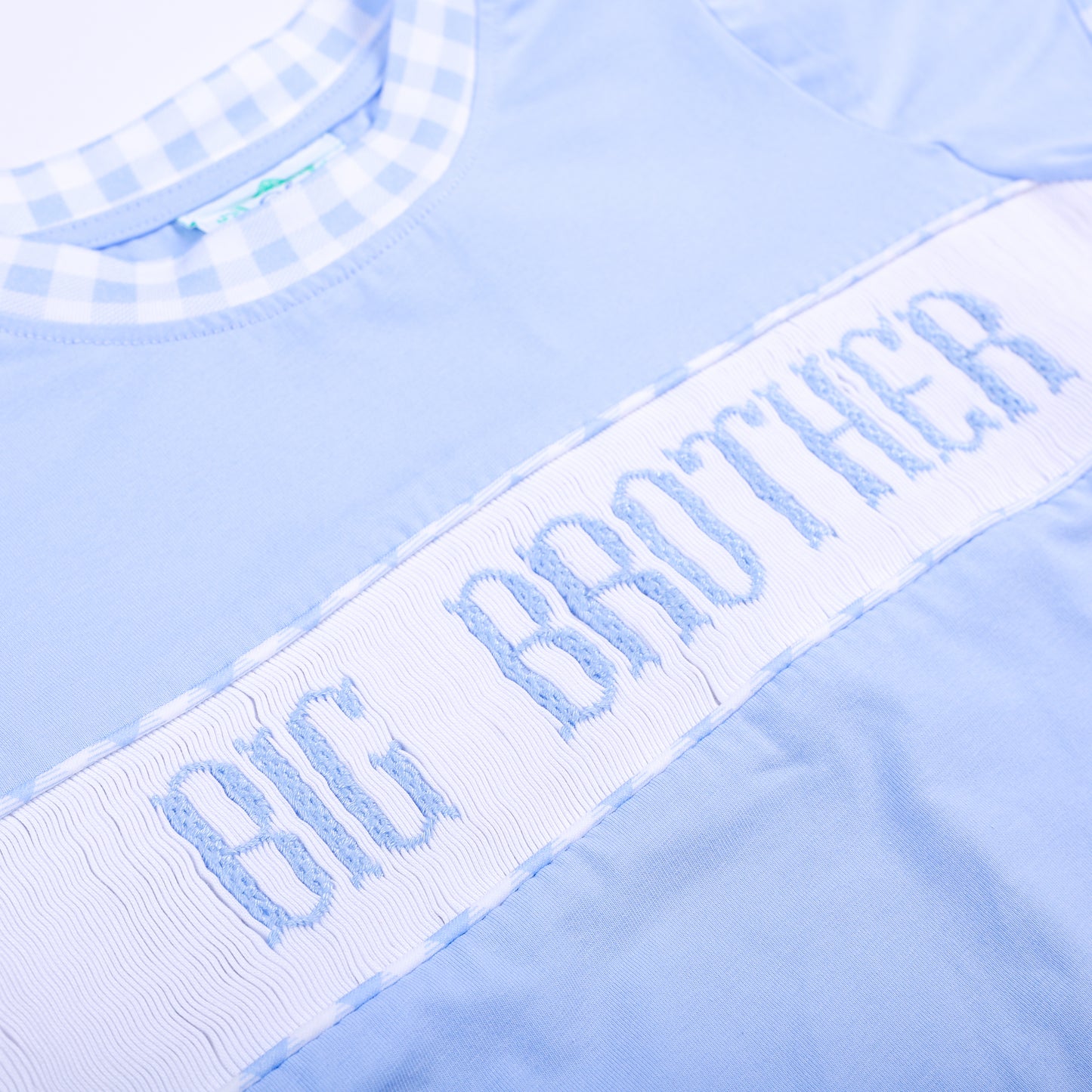 Angled close-up of delicate hand smocked and embroidered Big Brother lettering on light blue cotton shirt.