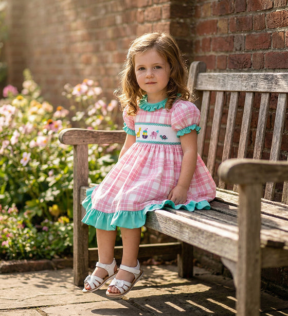 Little girl in a pink plaid smocked dress with teal ruffle trim and embroidered animals sitting on a bench.