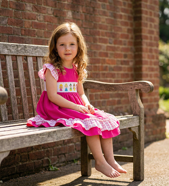 Little girl in a hot pink smocked princess dress with gingham flutter sleeves sitting on a wooden garden bench.