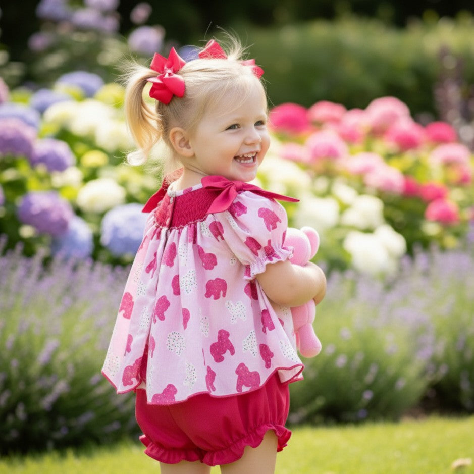Back view of toddler wearing hand smocked frosted sprinkle animal cookies set in a flower garden holding plush toy.