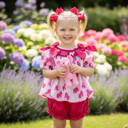 Toddler girl wearing hand smocked frosted sprinkle animal cookies set in a flower garden with a pink plush toy.