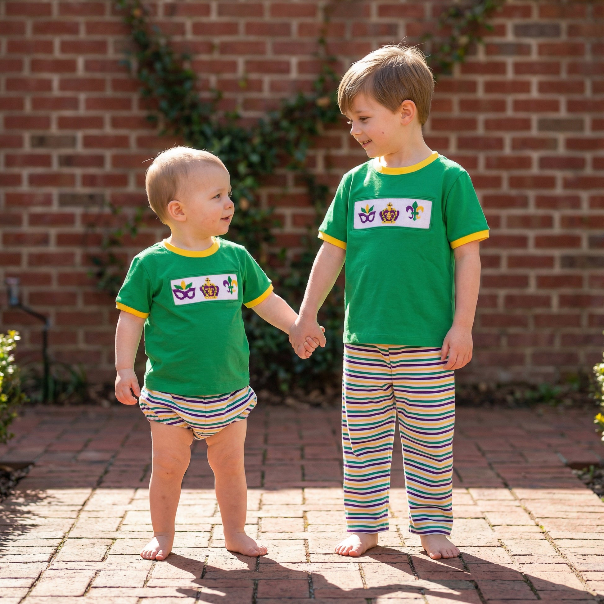 Brothers holding hands in green hand smocked Mardi Gras pant set and matching bloomers with striped bottoms.