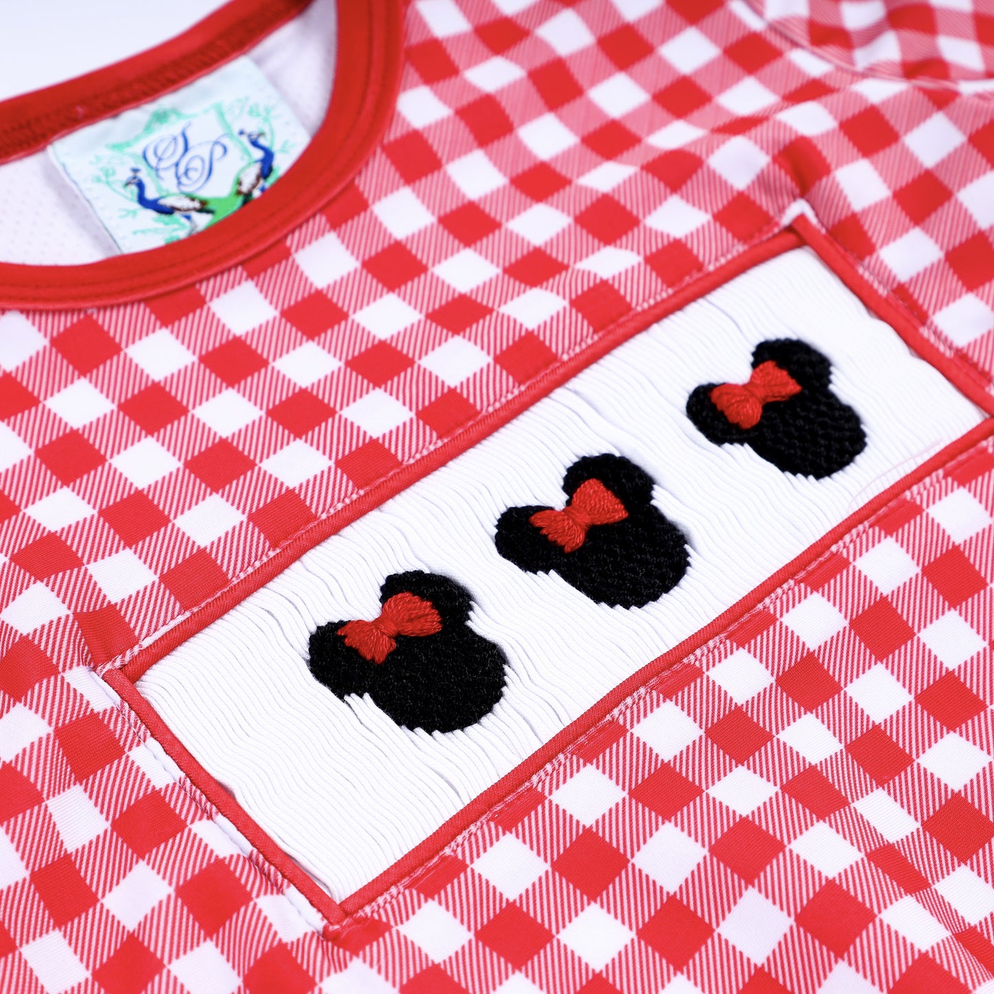 Detailed macro view of hand embroidered Minnie Mouse icons on white hand smocked chest plate of red gingham swimsuit.