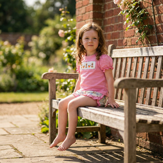 Girl on bench wearing pink Peter Rabbit hand smocked top and shorts set in a garden.