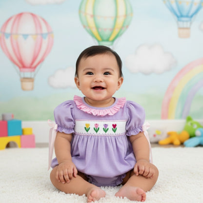 Smiling baby girl sitting on white rug wearing a hand smocked purple swiss dot tulip bubble outfit with ribbons.