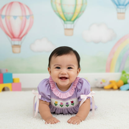 Baby lying on tummy wearing a hand smocked lavender swiss dot tulip bubble outfit with pink ruffle collar.