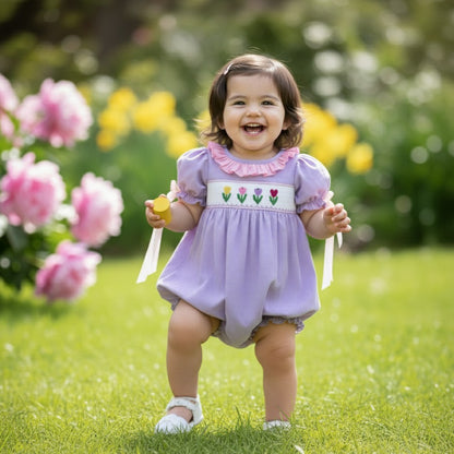 Toddler standing on grass wearing a hand smocked purple swiss dot tulip bubble with embroidered flowers.