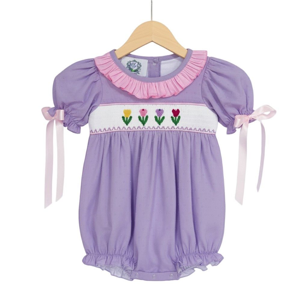 Hand smocked lavender swiss dot tulip bubble outfit for baby girls displayed on a wooden clothing hanger.
