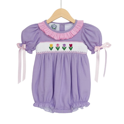 Hand smocked lavender swiss dot tulip bubble outfit for baby girls displayed on a wooden clothing hanger.
