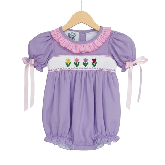 Hand smocked lavender swiss dot tulip bubble outfit for baby girls displayed on a wooden clothing hanger.