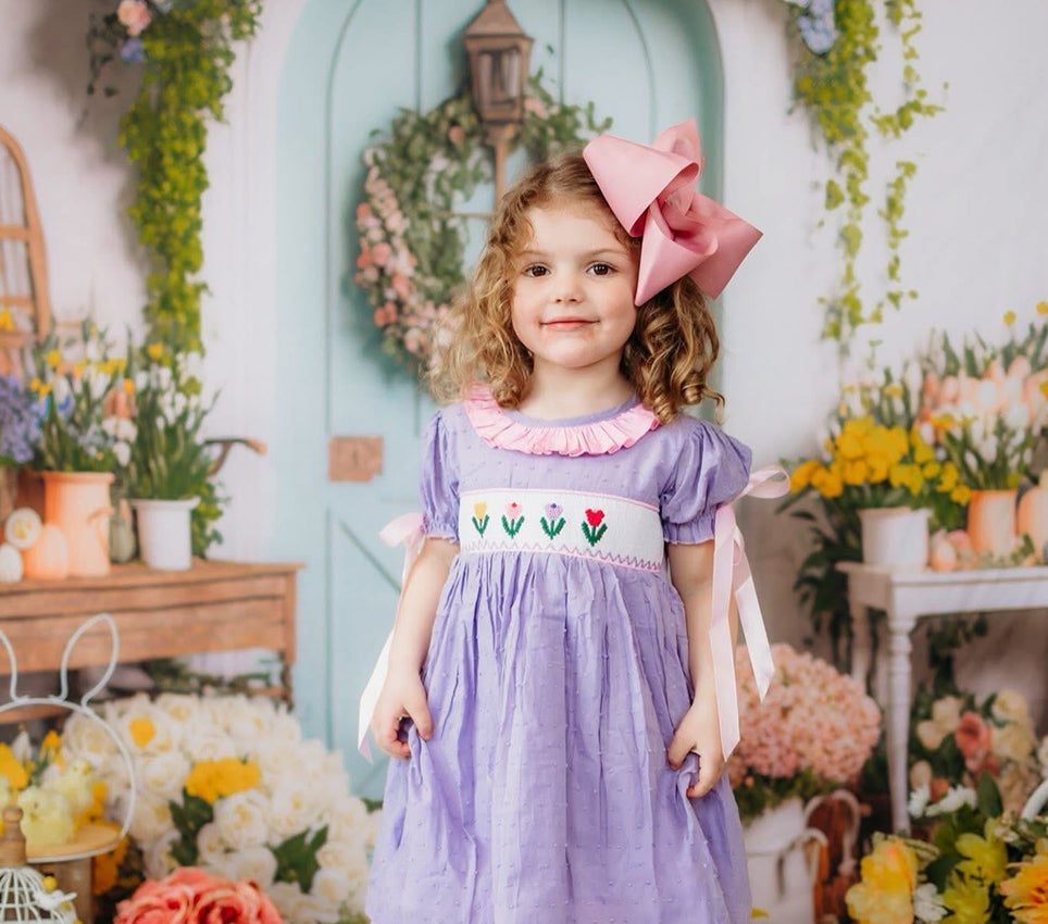Little girl wearing a hand-smocked swiss dot tulips dress in lavender with pink ruffle collar and hair bow.