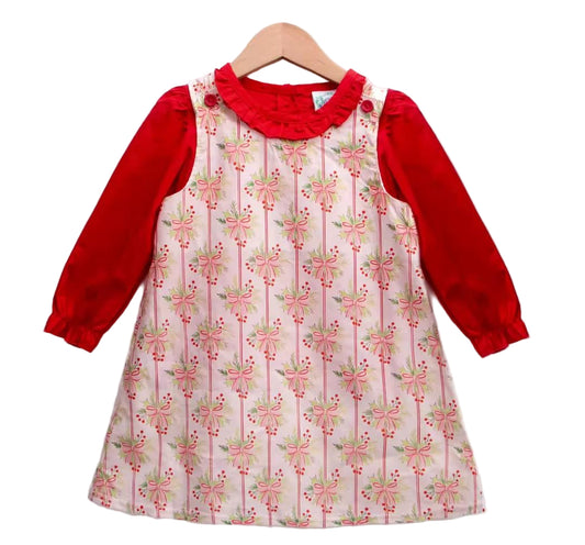 Product shot: Red long-sleeve ruffled blouse under a woven cotton jumper with holly berry and pink bow print.